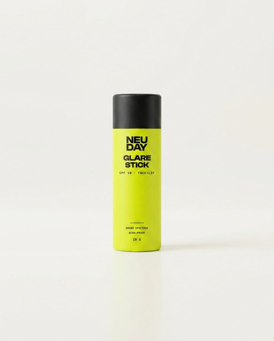 NEUDAY Glare Stick SPF 50 — a volt-yellow face and lip sunscreen stick with a matte-black base, on a warm bone studio background